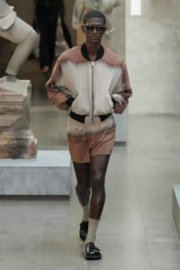 Dark-skinned model walks a runway wearing a beige-brown gradient bomber jacket and matching shorts, with sunglasses on.