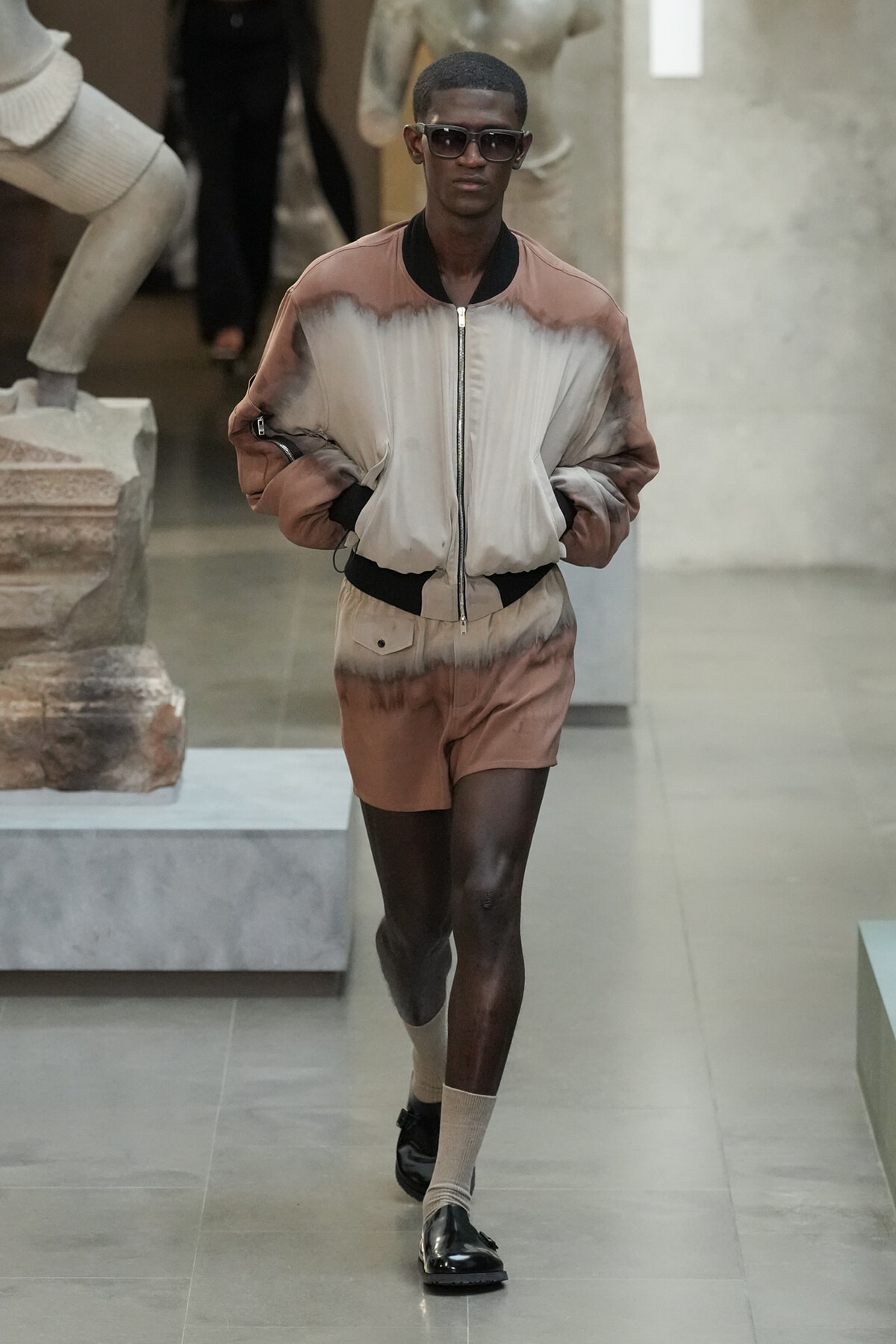 Dark-skinned model walks a runway wearing a beige-brown gradient bomber jacket and matching shorts, with sunglasses on.