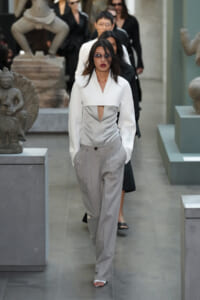 Model walks a runway-like corridor in a museum wearing a white structured jacket with a front cutout and light gray pleated trousers, sunglasses on.