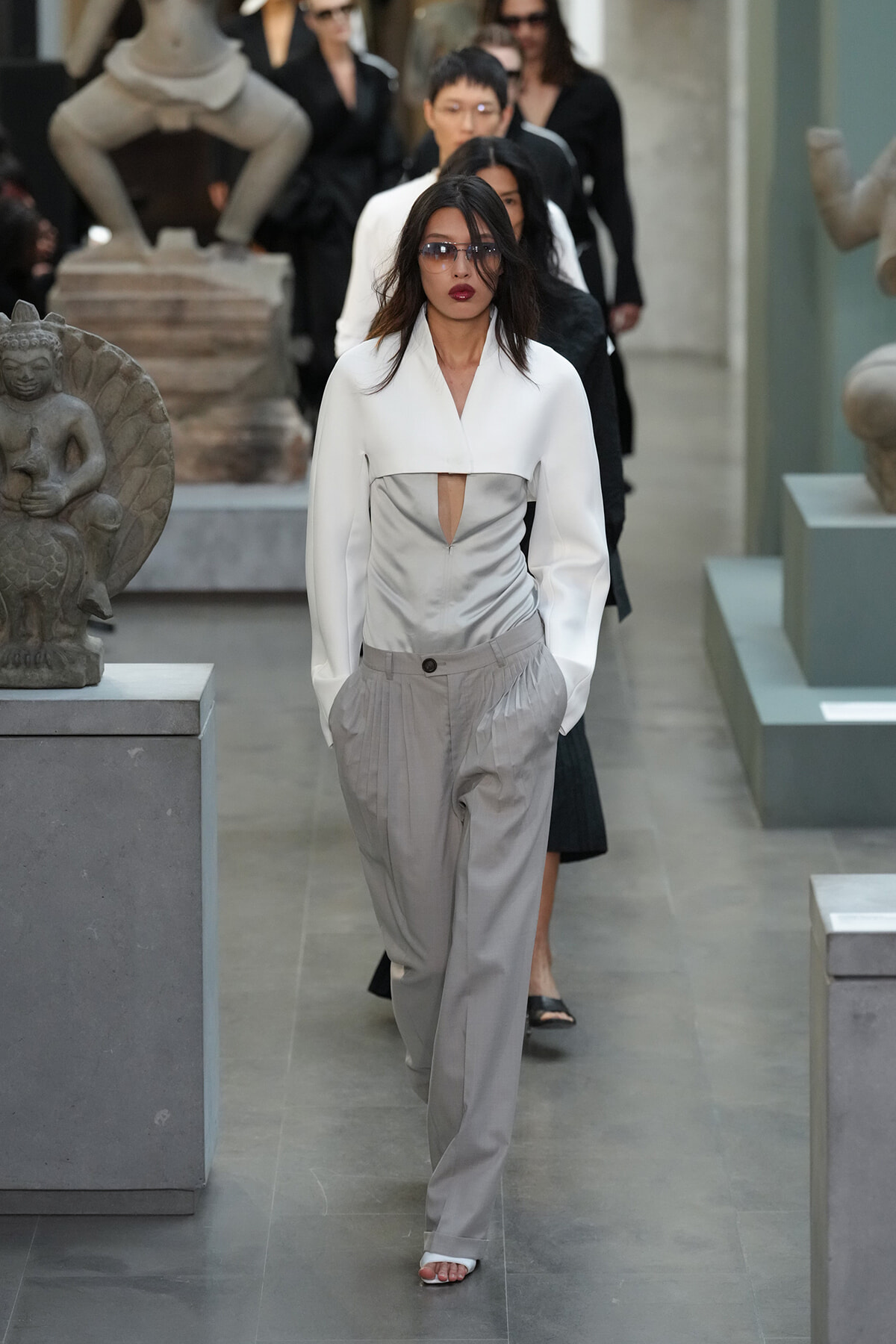 Model walks a runway-like corridor in a museum wearing a white structured jacket with a front cutout and light gray pleated trousers, sunglasses on.