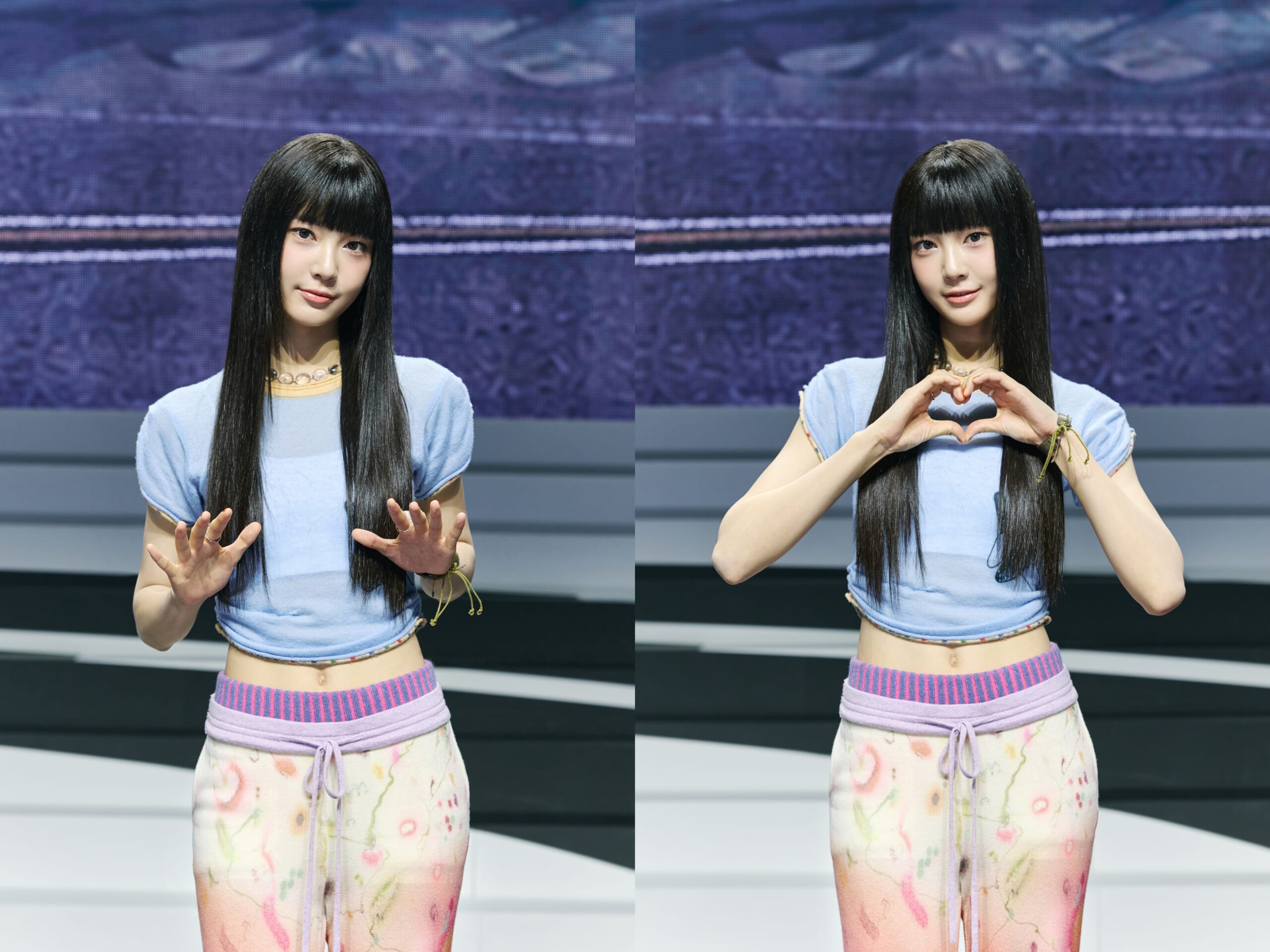 Two women on a stage wearing light blue tops; left figure raises hands palm-out, right figure forms a heart with hands against a blue backdrop.
