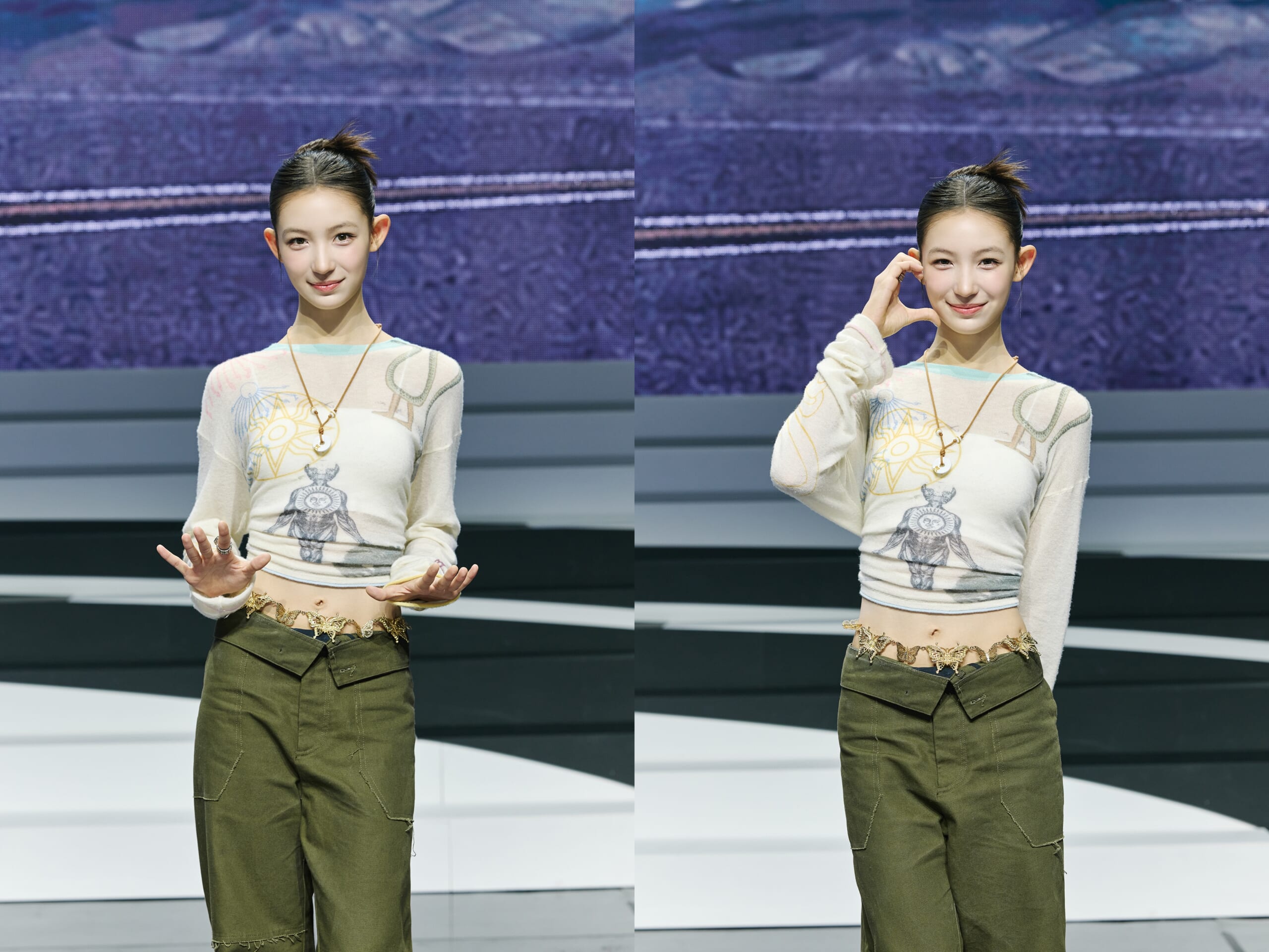 Young woman on stage wearing a graphic sweater and olive cargo pants, gesturing with open hands in front of a blue backdrop.
