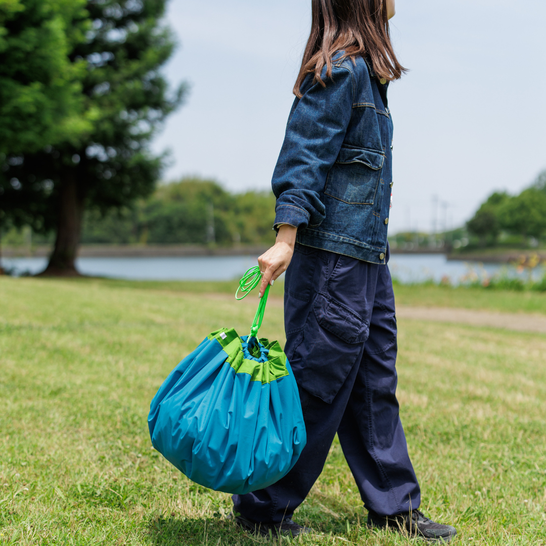 Person walking on grass beside a lake, carrying a bright blue and lime-green drawstring bag by the handle.