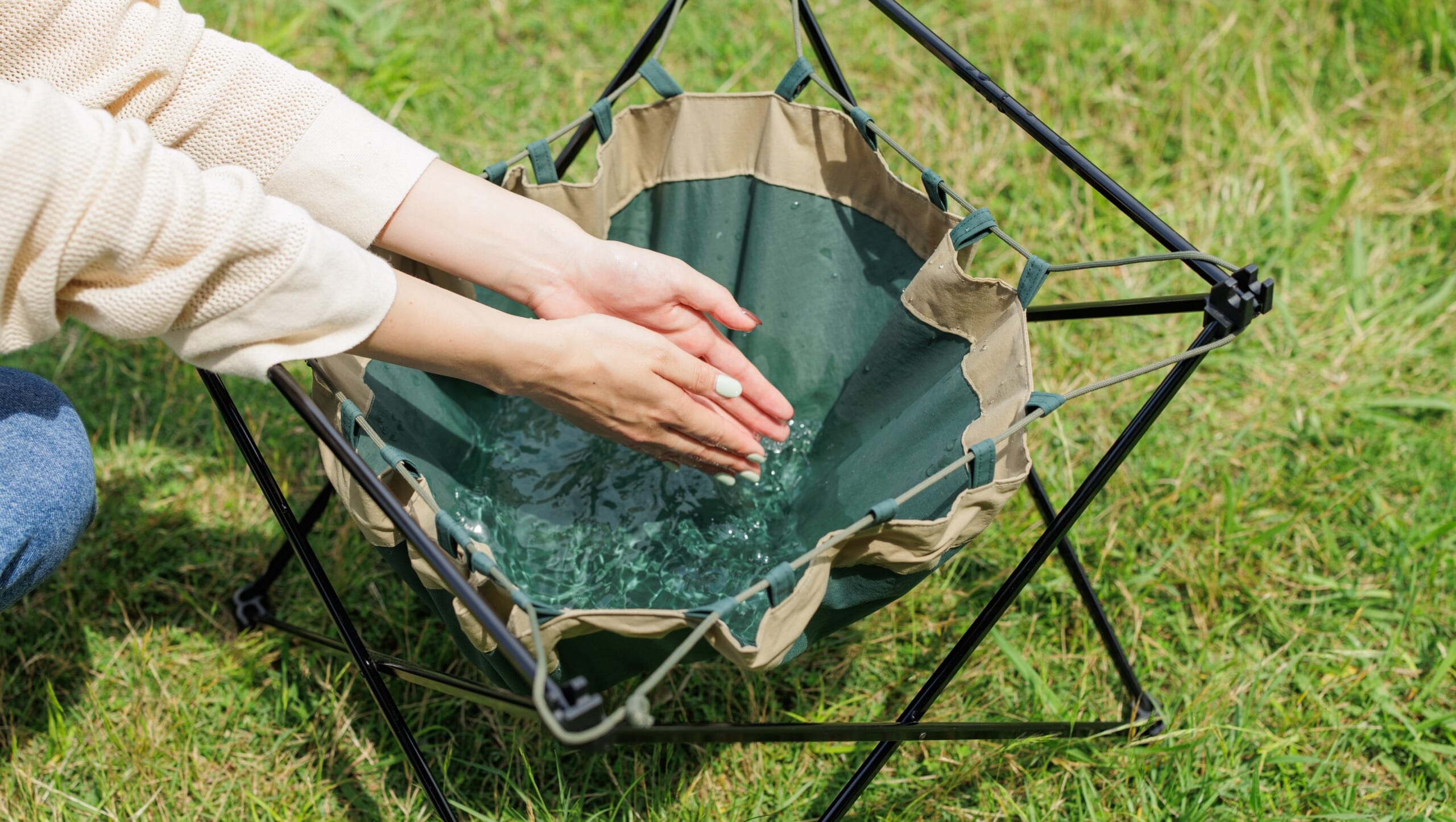 Hands washing under running water in a green fabric bucket suspended in a metal frame outdoors on a grassy surface, close-up view.