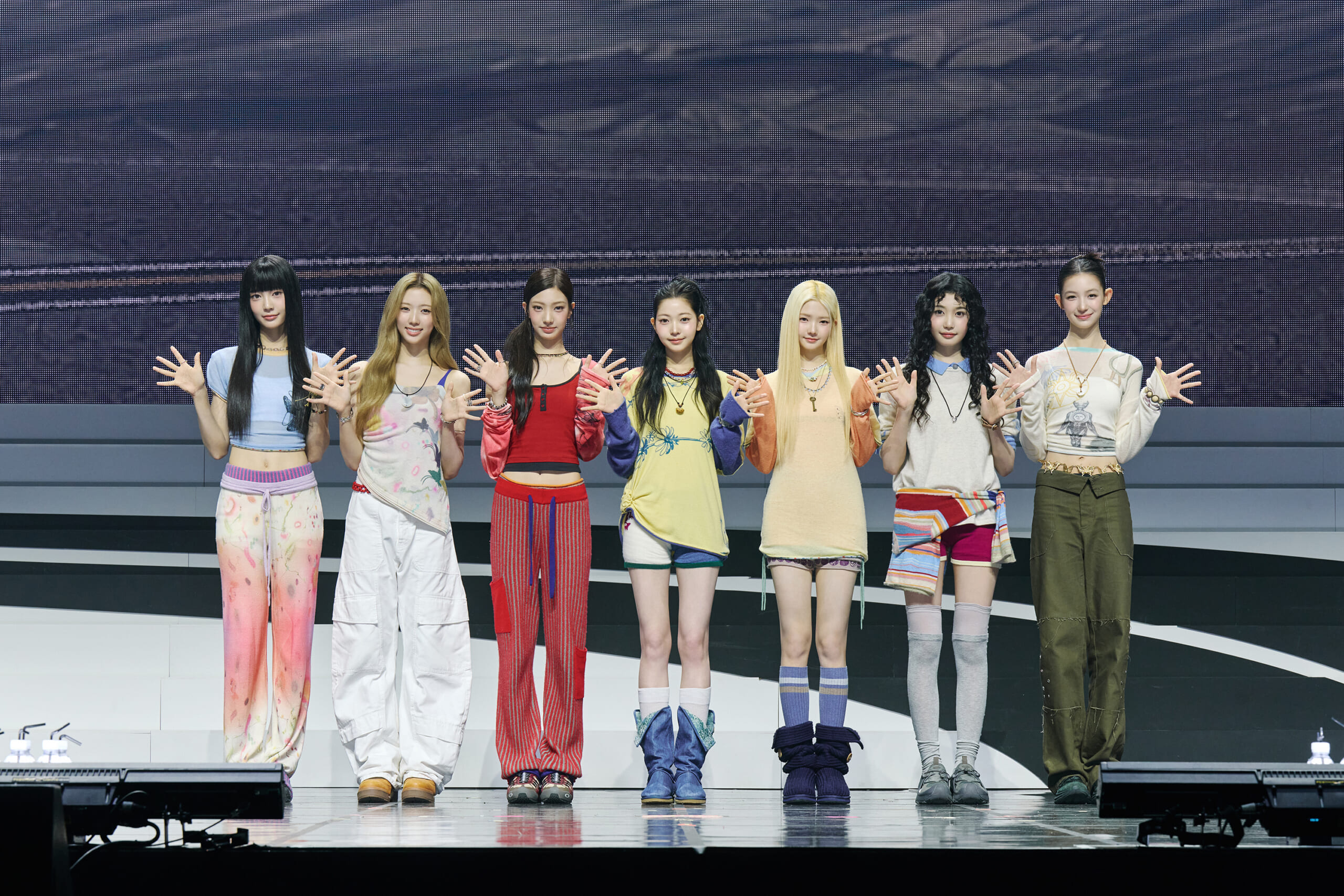 Seven young women in colorful outfits stand on a tiered stage, waving at the audience with smiles.