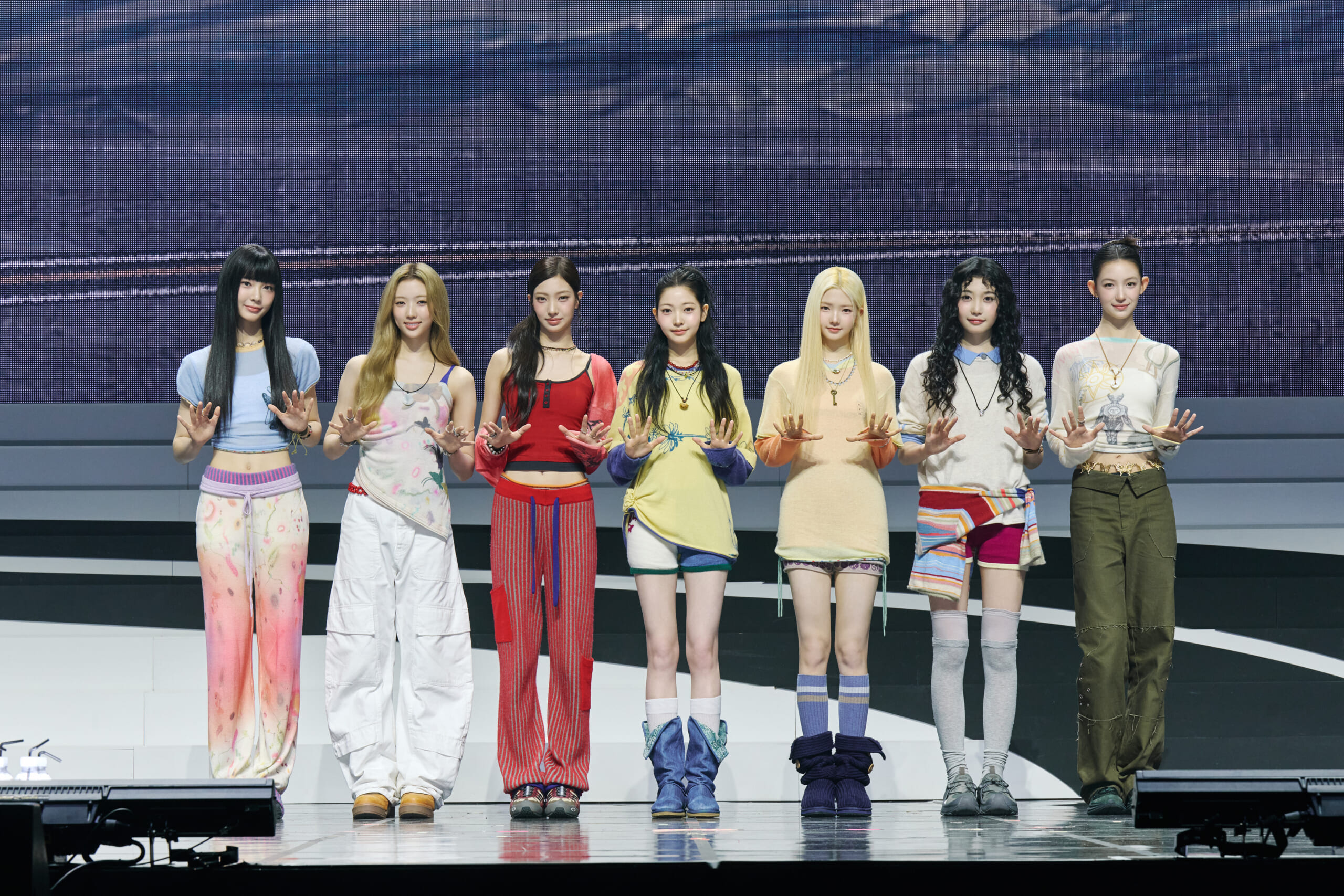 Seven women stand in a line on a stage, wearing colorful coordinated outfits and holding their hands forward toward the camera.